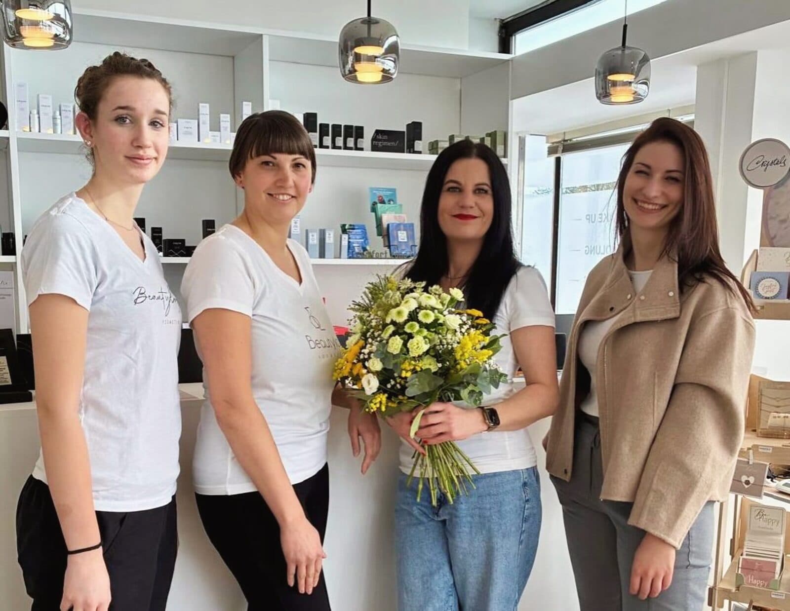 Das Beautyloft-Team im Institut: vier Kolleginnen nebeneinander vor dem Produktregal, eine mit Blumenstrauß